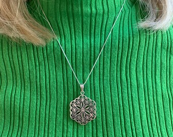 May include: A silver necklace with a geometric flower of life pendant. The necklace is on a person wearing a green turtleneck sweater.