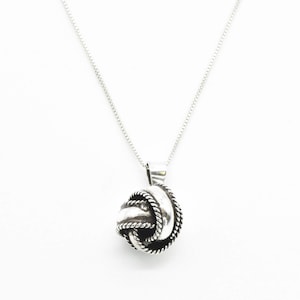 Puede incluir: Un collar de plata con un colgante en forma de nudo. El colgante está hecho de alambre de plata trenzado y tiene un acabado liso y pulido.