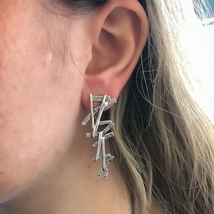 May include: Silver earrings with a geometric design. The earrings are made of multiple pieces of metal that are connected together to form a long, dangling shape.