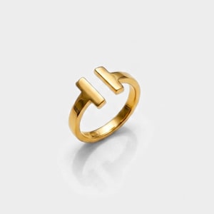 May include: A polished gold ring featuring an open design with two T-shaped ends that face each other. The band has a smooth, reflective surface and a consistent metallic yellow tone throughout the entire structure of the jewellery piece.