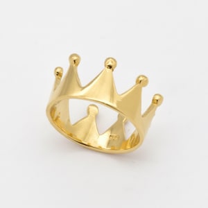 May include: A gold-colored crown ring with a detailed design. The ring features a series of pointed arches, each topped with a small, rounded orb. The ring is set against a plain white background.