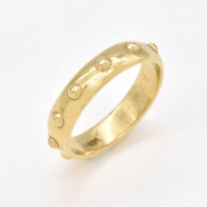 May include: A gold-colored ring with a series of small, raised circular details evenly spaced around the band. The ring has a smooth, polished surface and a classic, elegant design. The ring is likely made of gold or a gold-toned metal.