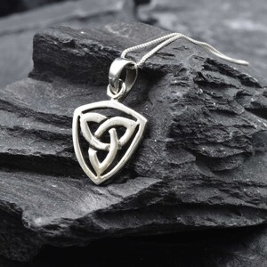 May include: A silver Celtic knot pendant in the shape of a triangle. The pendant is on a silver chain and is resting on a dark gray rock.