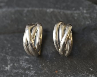May include: A pair of silver-toned hoop earrings. Each earring features a woven design of intertwined, curved metal bands. The earrings are displayed on a dark, textured surface, showcasing their polished finish and intricate details.