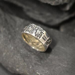 May include: A silver ring featuring an intricate design of stylised figures and symbols. The ring is wide and has a polished finish.
