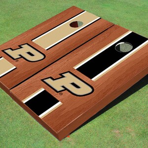May include: A wooden cornhole board set with a brown wood grain finish. The board features the letter "P" in black and gold, along with black and gold stripes. The board has a hole for bean bag toss.