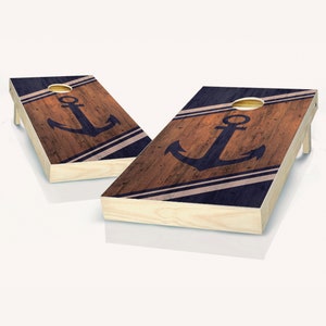 May include: Two wooden cornhole boards with a navy blue anchor design on a brown woodgrain background. The boards have white stripes and a distressed look.