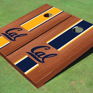May include: A wooden cornhole board set with a brown finish. Each board features a navy blue and gold stripe design with the word "Cal" in navy blue. The boards have a hole for tossing bean bags.