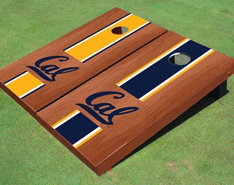 Op de afbeelding: Een houten cornhole-bordenset met een bruine afwerking. Elk bord heeft een marineblauw en goudkleurig streepontwerp met het woord "Cal" in marineblauw. De borden hebben een gat om zakken te gooien.