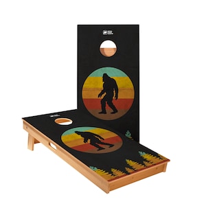 May include: Cornhole game set featuring a black surface with a Bigfoot silhouette inside a retro sunset circle. The set includes two wooden boards with a hole and a natural wood frame. The design also includes a forest scene.