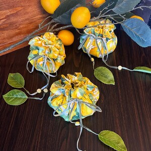 three lemon bags on a wooden backdrop. each bag is closed showing a lemon patterned fabric and a drawstring tied in a bow with leaves and lemon slice charms attached to the ends.