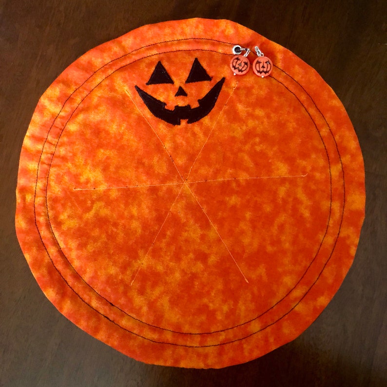 Puede incluir: Un tapete redondo de tela naranja con textura moteada que presenta una cara de calabaza bordada en negro. El tapete tiene l&iacute;neas de costura radiales y dos peque&ntilde;os dijes circulares de calabaza sujetos al borde superior con anillas de metal plateado.