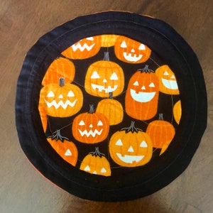 Puede incluir: Un cuenco de tela acolchada redondo con un fondo negro y un patr&oacute;n repetitivo de calabazas naranjas con caras talladas en blanco. El art&iacute;culo circular tiene un borde ancho de tela negra s&oacute;lida cosido alrededor del borde exterior.