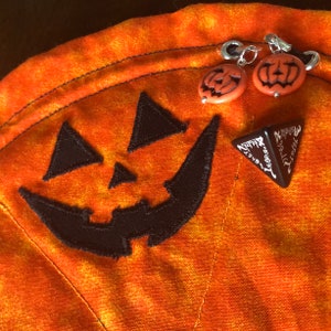 Puede incluir: Una bolsa de tela naranja con una cara de calabaza bordada en negro. Dos peque&ntilde;os dijes en forma de calabaza naranja est&aacute;n unidos al tirador de cremallera plateado. Un dado triangular marr&oacute;n oscuro con texto blanco sobre la tela.