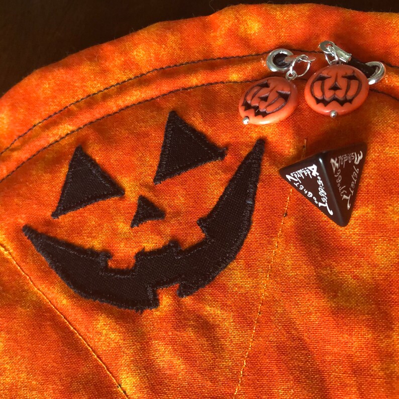 Puede incluir: Una bolsa de tela naranja con una cara de calabaza bordada en negro. Dos peque&ntilde;os dijes en forma de calabaza naranja est&aacute;n unidos al tirador de cremallera plateado. Un dado triangular marr&oacute;n oscuro con texto blanco sobre la tela.