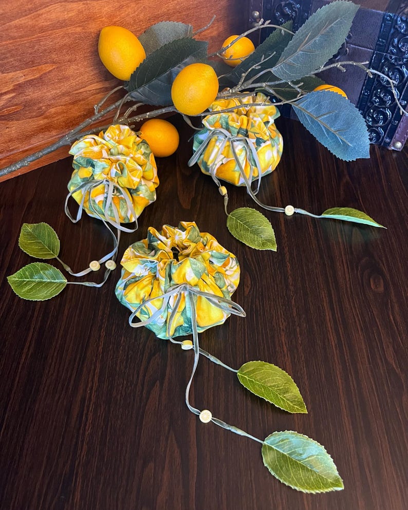 three lemon bags on a wooden backdrop. each bag is closed showing a lemon patterned fabric and a drawstring tied in a bow with leaves and lemon slice charms attached to the ends.