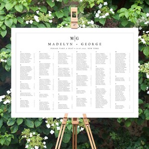 May include: A white wedding seating chart with the names Madelyn and George printed at the top. The chart is on an easel and lists the names of guests and their assigned tables.