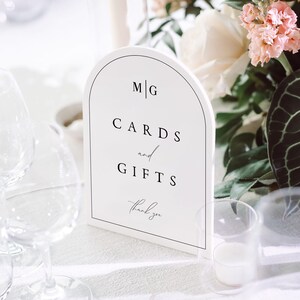 May include: White card with the text "M|G CARDS and GIFTS thank you" on a white table with a bouquet of pink and white flowers, a candle, and glasses.