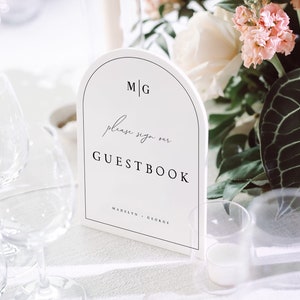 May include: White sign with a rounded arch top, reading "M|G please sign our GUESTBOOK Madelyn & George". The sign is on a white tablecloth with two clear wine glasses and a vase of pink and white flowers.