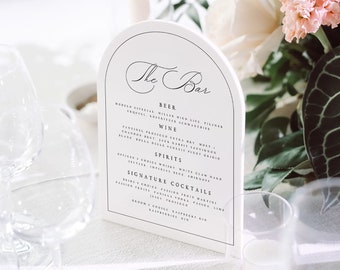 May include: A wedding reception table setting with a floral arrangement of pink and white roses, a clear glass candle holder with a white candle, and a menu card that reads "The Bar". Several wine glasses are in the foreground.