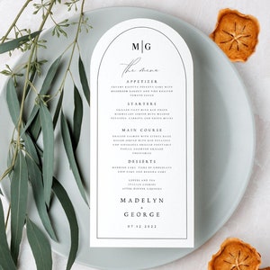 May include: A white wedding menu card with a black arch design and the text "M|G The Menu" at the top. The menu includes appetizers, starters, main course, desserts, coffee and tea, and the names Madelyn & George with the date 07.12.2022.