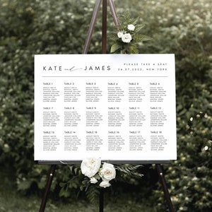 May include: A white wedding seating chart on an easel with the names "Kate and James" and the date "24.07.2022, New York" printed at the top. The chart lists the names of guests assigned to each table number.