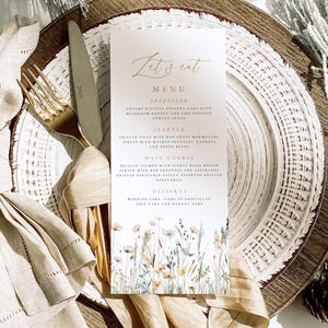 May include: A formal table setting with a menu card. The menu card has the text "Let's eat" in gold script. The card features a floral design at the bottom. Gold cutlery and a beige napkin are also present.