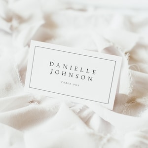 Puede incluir: Tarjeta blanca con texto negro que dice "Danielle Johnson Mesa Uno". La tarjeta está sobre una superficie de tela blanca.