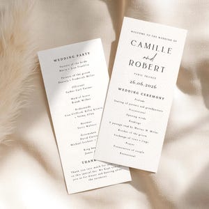 Può includere: Due eleganti programmi della cerimonia nuziale su sfondo crema. I programmi presentano testo nero con i nomi "Camille e Robert" e la data "26.06.2026". Un programma elenca gli invitati al matrimonio, l'altro i dettagli della cerimonia.