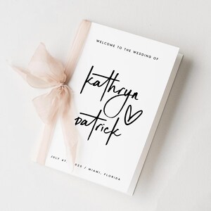 May include: A white wedding program with the text "WELCOME TO THE WEDDING OF Kathryn Patrick" in black script. A blush-coloured ribbon is tied around the program. The date "JULY 4T" and location "020 / MIAMI, FLORIDA" are also printed.