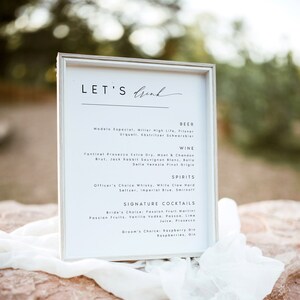 May include: A white framed sign with the text "LET'S drink" at the top. The sign lists a variety of beer, wine, spirits, and signature cocktails.