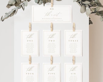 May include: White cardstock table number cards with gold calligraphy. The cards are hung on a white board with wooden clothespins. The cards are numbered one through six and each card has a list of names.
