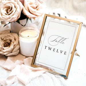 May include: Gold framed table number card with the text "Table Twelve" in a cursive font. The card is surrounded by white roses, a white candle, and a white ribbon.