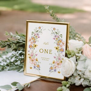 Puede incluir: Una tarjeta con número de mesa con marco dorado y detalles florales. La tarjeta dice "table ONE" con los nombres "Sarah & James" y la fecha "26.06.20xx". La tarjeta está rodeada de vegetación y flores.