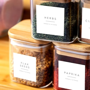 May include: Four glass jars with bamboo lids and white labels. The labels have black text that reads "Herbs", "Flax Seeds", "Paprika", and "Cinnamon". Each label also includes the text "Sarah's Kitchen".
