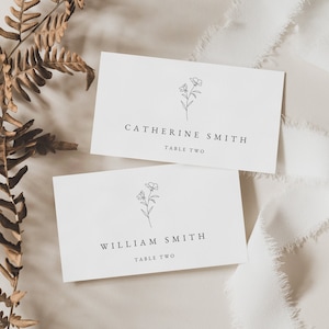 Puede incluir: Dos tarjetas de lugar blancas con un diseño floral sencillo y los nombres "Catherine Smith" y "William Smith" con el número de mesa "Mesa Dos" impreso en cada una.