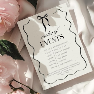 May include: A wedding schedule card with a black bow and decorative border. The card lists the timings for the wedding ceremony, cocktail hour, dinner, and dancing. Pink flowers are arranged around the card.
