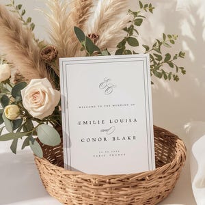 Op de afbeelding: Een trouwuitnodiging in een rieten mand, omringd door bloemen en pampasgras. De kaart leest "Welkom bij de bruiloft van Emilie Louisa en Conor Blake", met de datum en locatie, Parijs, Frankrijk.