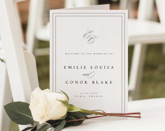 May include: A white wedding program with black text, resting on a white chair. The program reads "Welcome to the Wedding of Emilie Louisa and Conor Blake" with the date and location. A white rose and greenery are placed on the chair.