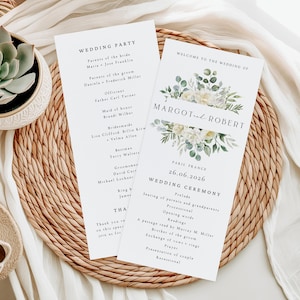 May include: Two white wedding programs with floral designs and the names "Margot and Robert" are displayed on a woven placemat. The program includes the wedding date, location, and ceremony details.