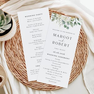 Könnte beinhalten: Zwei weiße Hochzeitskarten mit schwarzem Text, eine mit "Wedding Party" und die andere mit "Welcome to the Wedding of Margot and Robert". Beide Karten sind mit floralen Designs verziert und liegen auf einem gewebten Platzset.
