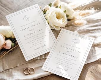 Op de afbeelding: Twee trouwuitnodigingen met zwarte tekst en een dubbele rand, naast gouden trouwringen en witte bloemen. De uitnodigingen luiden "Emilie Louisa en Conor Blake" met evenementdetails. De achtergrond is een lichtgekleurde stof.