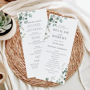 May include: Two white wedding ceremony programs with eucalyptus leaf accents. One program lists the wedding party, and the other welcomes guests to Delilah and Robert's wedding in Paris, France, on June 26, 2026.