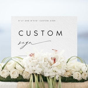 May include: A white sign with black text that reads "5"x"7 and 8"x"10" Custom Sign" and "CUSTOM sign" in cursive. The sign is sitting on a table with white flowers.