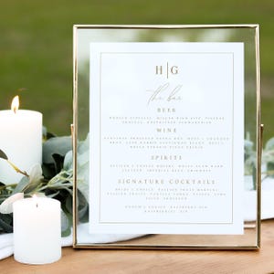 May include: A gold-framed sign with a white background and gold lettering lists the bar menu for a wedding reception. The menu includes beer, wine, spirits, and signature cocktails. The couple's initials, "H|G", are at the top of the sign.