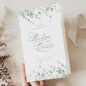 Könnte beinhalten: Ein weißes Hochzeits-Willkommensschild mit dem Text "Willkommen zur Hochzeit von Madison und Quentin" in eleganter Schrift. Das Schild ist mit Aquarell-Eukalyptusgrün geschmückt. Datum und Ort sind ebenfalls angegeben.