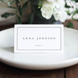 Puede incluir: Una tarjeta de lugar blanca con un borde negro y el texto "ANNA JOHNSON" y "TABLE 5" impreso en ella.