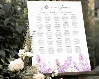 Op de afbeelding: Een witte trouwzittingstafel met de namen "Alice and James" in sierlijke letters. De tabel bevat tafeltoewijzingen en is versierd met aquarel lavendel en witte bloemen. Het bord wordt gepresenteerd op een houten ezel.