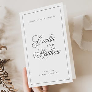 Può includere: Un invito di nozze bianco con testo nero. L'invito recita "Benvenuti al matrimonio di Cecilia e Matthew" con la data e il luogo sottostanti. L'invito è tenuto in mano.