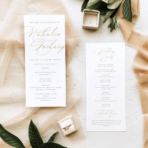 May include: Elegant wedding stationery featuring gold script on white paper. The design includes a wedding ceremony program and a wedding party list. A ring box and a ring are also visible, along with decorative fabric and greenery.
